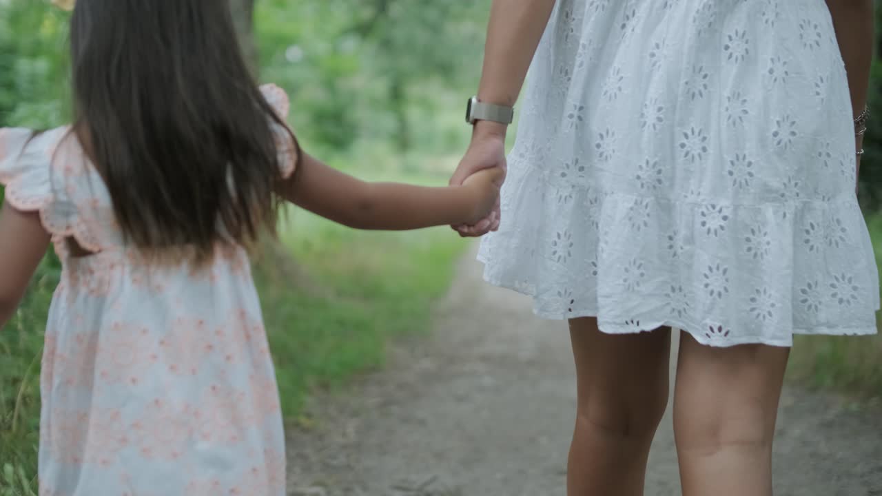 una mujer embarazada y su pequeña hija, ambas vestidas de blanco, caminan de la mano a través de un parque cubierto de hierba. la escena captura un momento sereno de unión familiar en la naturaleza, rodeada de árboles.