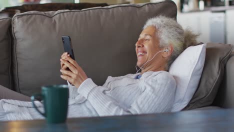 Senior-mixed-race-woman-lying-on-sofa-wearing-earphones-and-using-smartphone