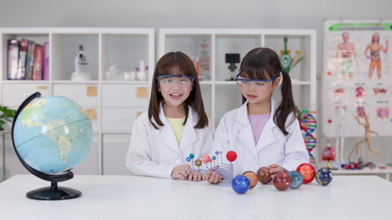 Two young girls in safety goggles examine a planetary model in a bright school laboratory.