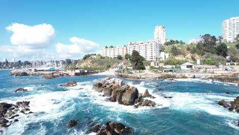 Olas-Del-Mar-Golpeando-Las-Rocas-Costeras-En-Viña-Del-Mar-Con-Edificios-Y-Puerto-Deportivo-Al-Fondo,-Chile