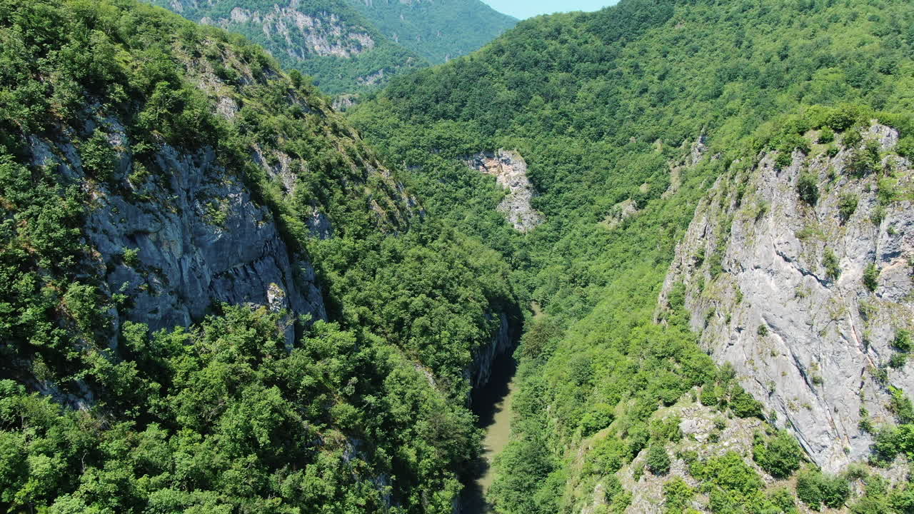 A view from above shows a river flowing through a valley. Tall cliffs line both sides of the river. The cliffs are covered in trees and other green plants. Mountains are visible in the background