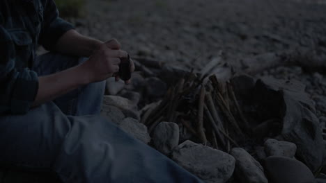 Medium-view-of-man-striking-match-to-start-campfire-tipi-in-fire-pit-on-pebble-beach,-slow-motion