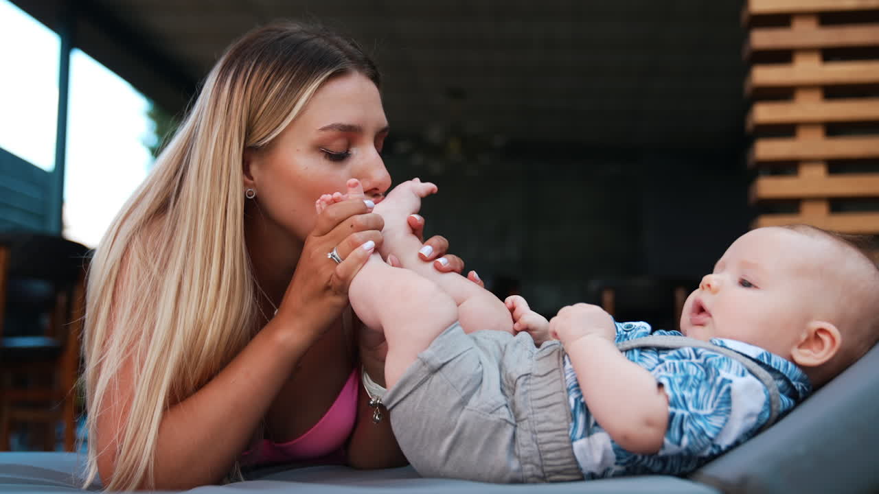 Loving mom kissing her infant son's tiny feet. Mother spending time with her child outdoors.