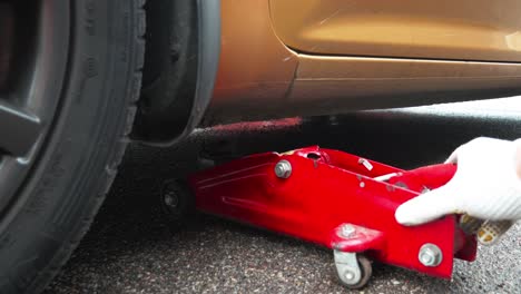 man hands pushing a red car jack under gold color car for wheel replacement