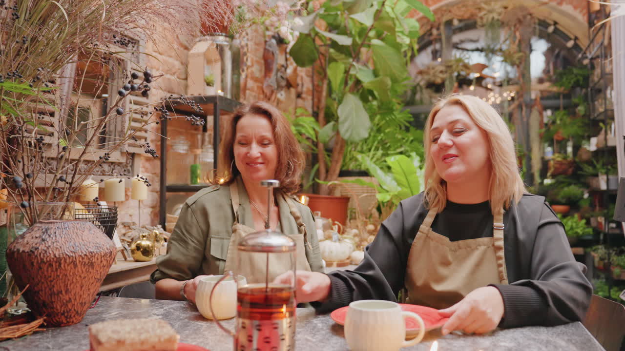 Floristas blancos degustando té de hierbas juntos; un empleado vierte infusión de prensa en la mesa de la tienda de plantas, con velas, jarrón y trozo de pastel. Llevan delantales, hay risas y revuelven juguetonamente, en un ritual detrás del mostrador.