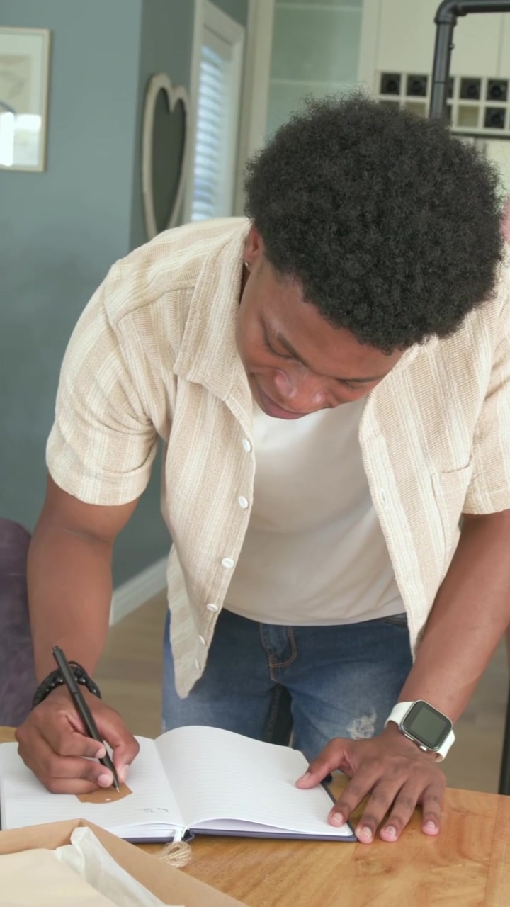 Vertical video: Leaning over notebook, man writing for work at home, reaching and placing sticker