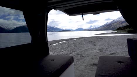 young traveler opening door and showing exterior and interior of motorhome by beautiful blue lake wakatipu, queenstown, new zealand with mountains fresh snow cloudy sky in background