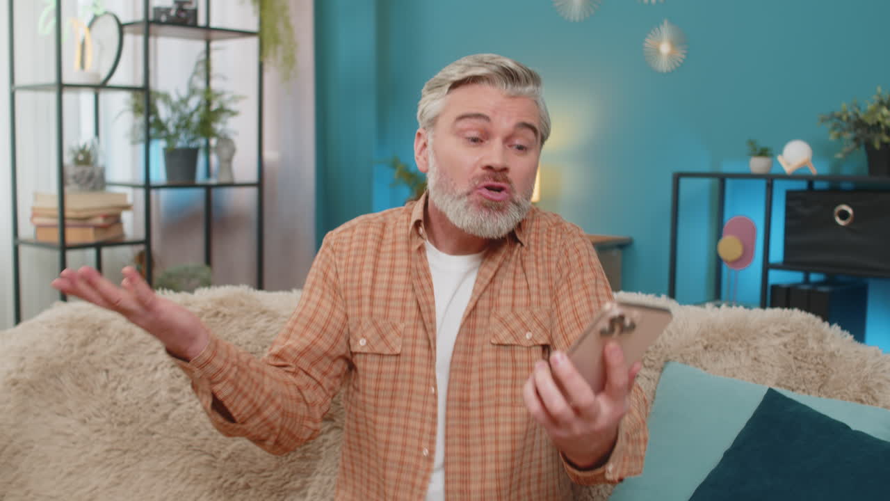 성숙한 남자가 집에서 소파에서 스트레스와 부정적인 감정을 표현하는 스피커에서 전화 논쟁을 하고 있습니다.