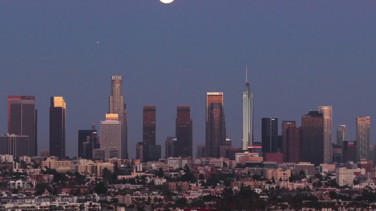 Telephoto tilting up aerial shot of the Los Angeles skyline under a full moon at magic hour in Southern California. 4K