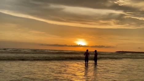 silhouette of two people facing the ocean on a beach with orange sunset at golden hour.