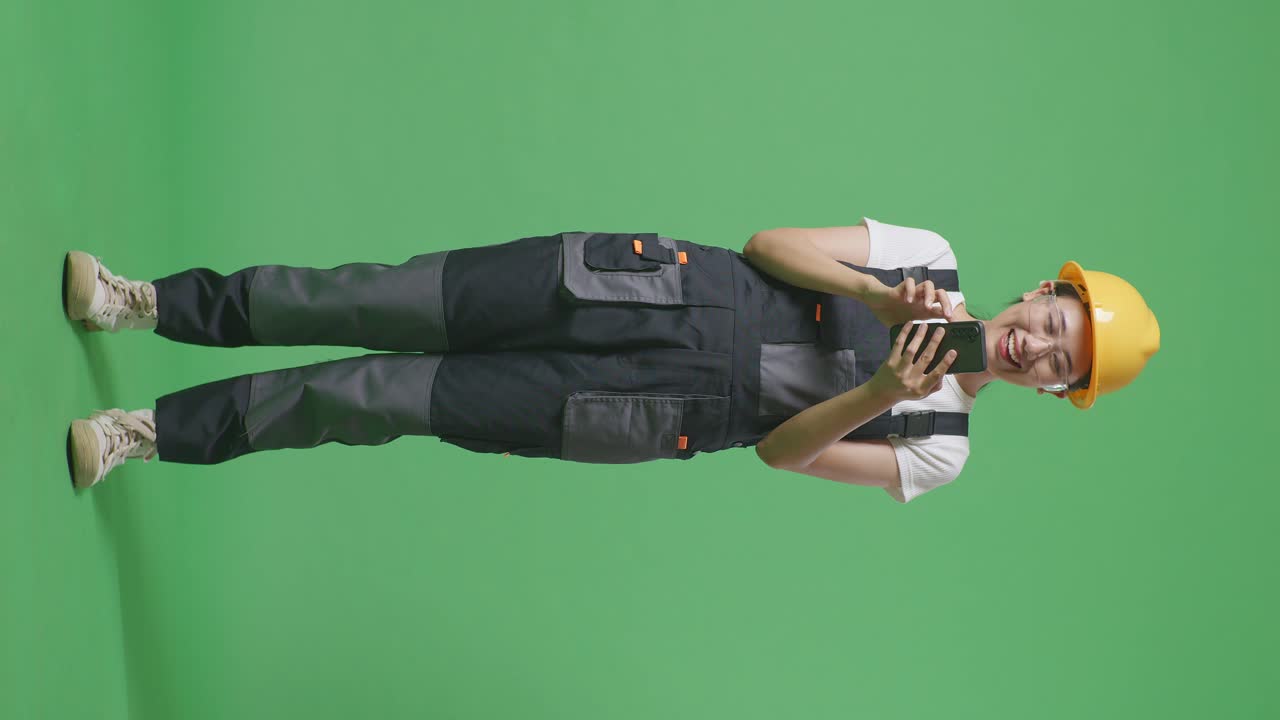 полное тело азиатской женщины-работницы в защитных очках и защитном шлеме улыбается и использует смартфон, стоя в фоновой студии с зеленым экраном