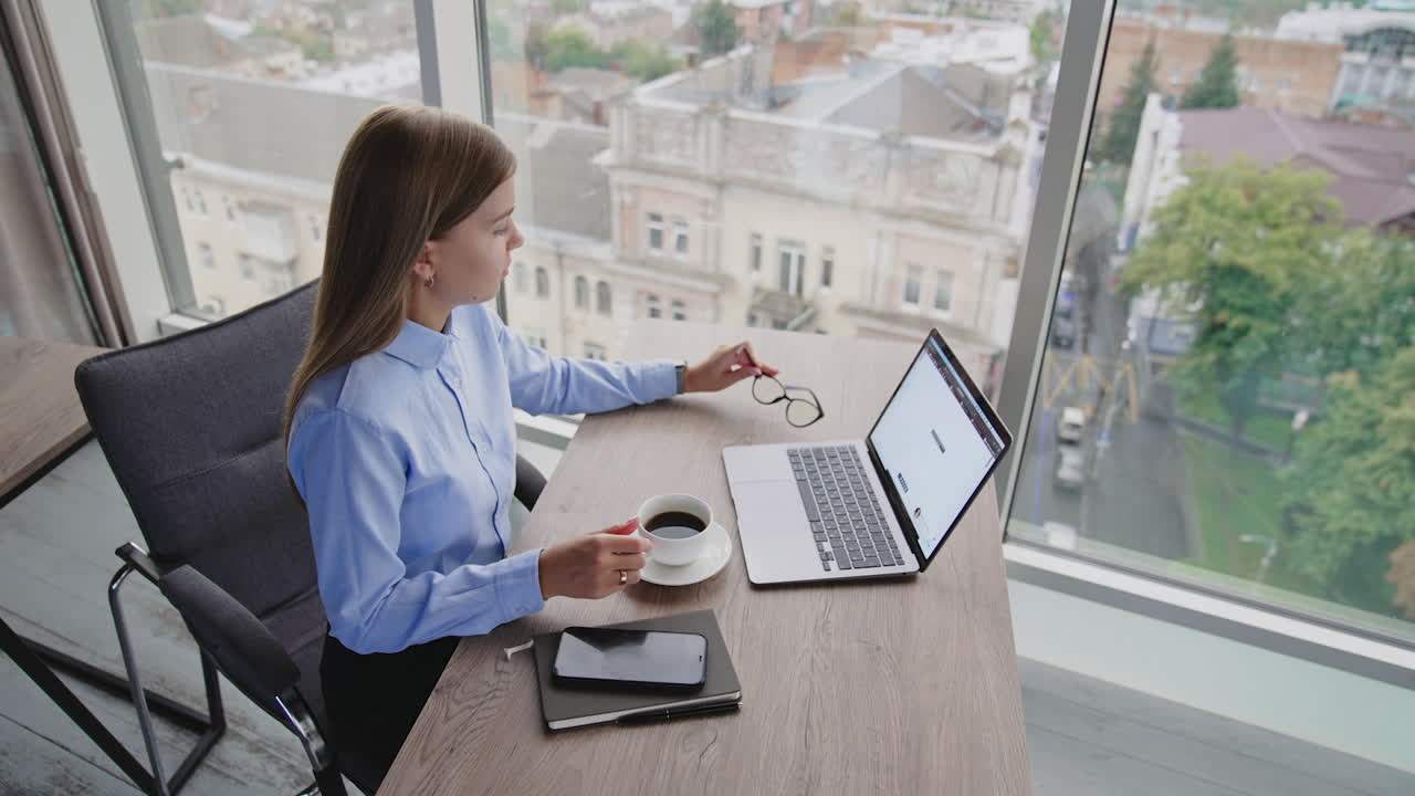 Busy young blonde lady working in the office having a break. Woman sitting in front of laptop takes a cup of coffee. High angle view.