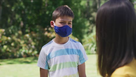 Portrait-of-caucasian-brother-and-sister-wearing-face-mask-standing-in-the-garden