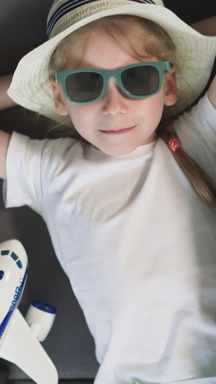 Blonde preschooler girl in sunglasses looks in camera lying on sofa near airplane and suitcase. Dreaming little girl ready for departure upper close view
