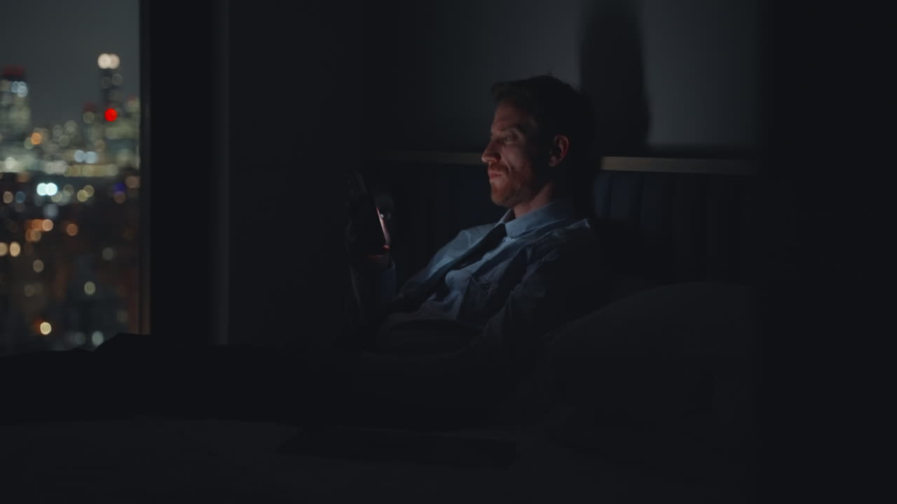 Bored businessman sitting in bedroom overlooking the city at night playing with smartphone or watching videos. Illuminated by phone screen he holds it and scrolls infinitely with tired emotionless