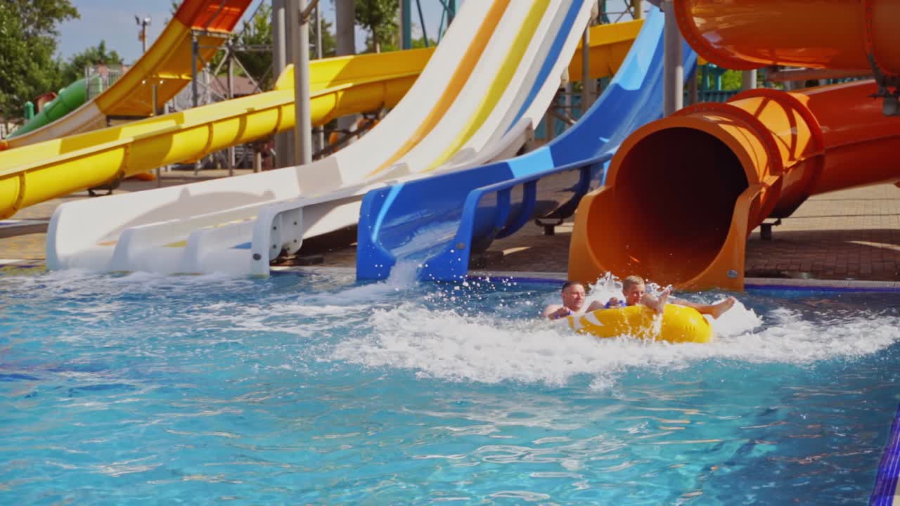 Father and son sliding from colorful pipe in the water park. Many slides in blue swimming pool in summer aquapark. Outdoor waterpark in amusement park. Joyful family vacation.