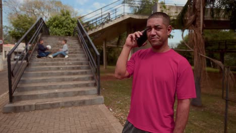 a man talking on a phone in an outdoor park with children playing on stairs in the background