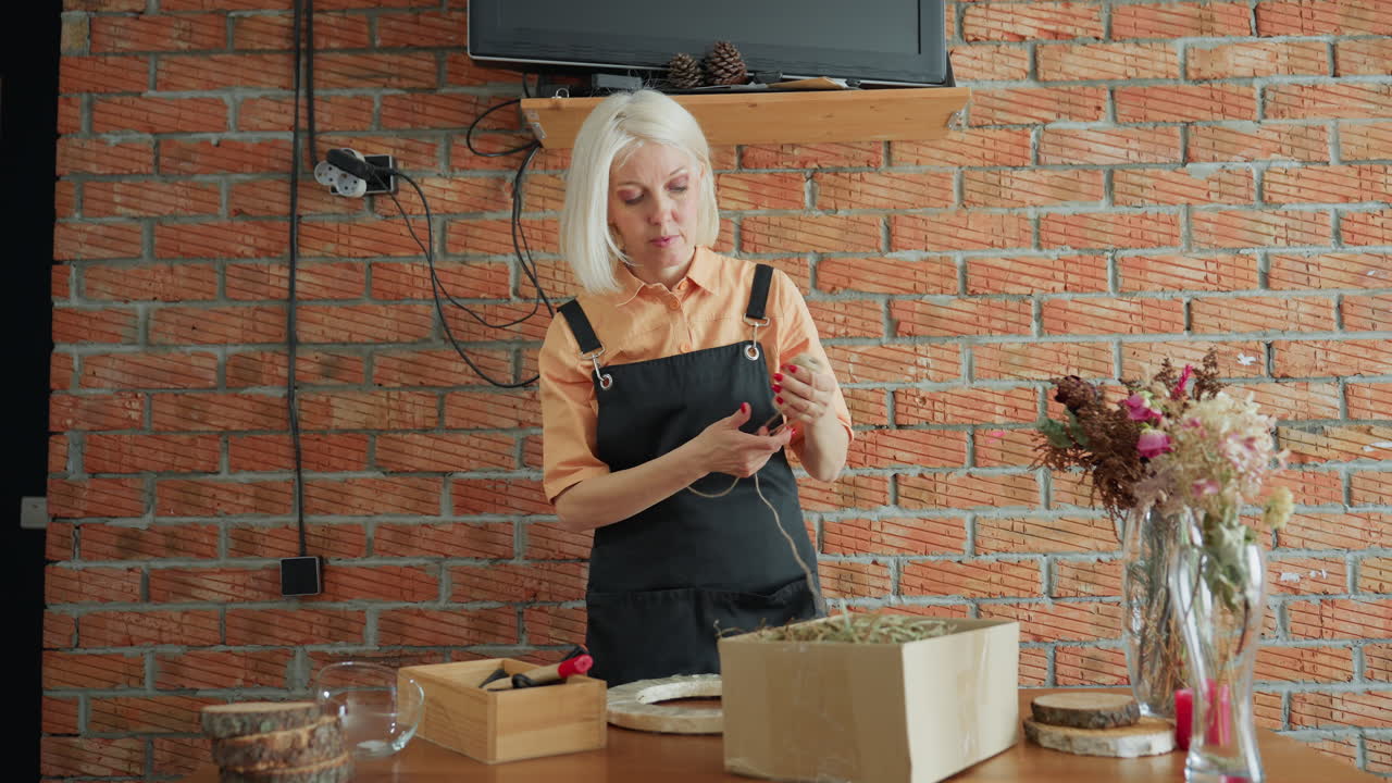 Florist in apron prepares handmade decoration with twine at wooden table, surrounded by dried flower arrangements, craft supplies, and natural elements against rustic brick wall interior in creative workspace