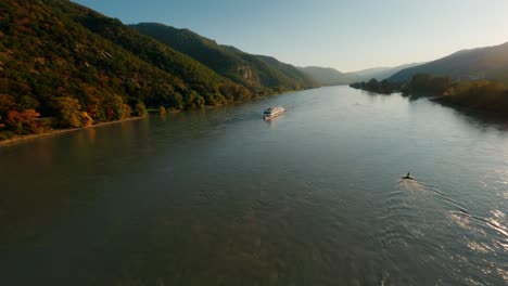 danube boat decorated with multiple flags approaching fpv drone, passing by at close proximity and making waves reflecting the beautiful sunset in wachau valley