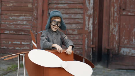 Cute-Little-Red-Haired-Boy-In-Hat-And-Glasses-Walking-Outdoors-In-A-Cardboard-Plane-Dreaming-To-Be-An-Aviator-1