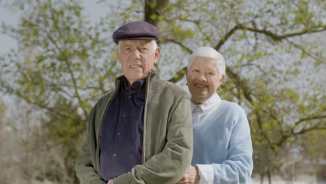 portrait shot of an elderly man looking at camera and smiling while his wife hugging him from behind on a sunny autumn day at park