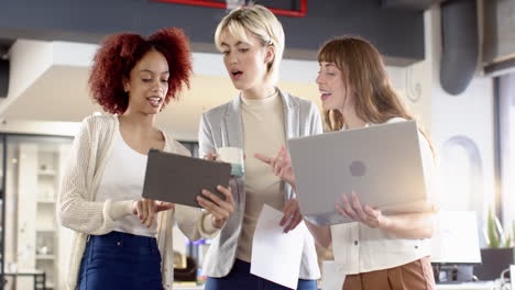discussing project, three women using tablet, laptop, and holding coffee in office