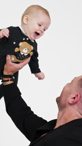 strong man holding his infant son with one hand. cute child in bodysuit with teddy smiles to dad adorably. white backdrop. vertical video