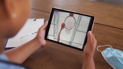 Composite-of-woman-sitting-at-table-with-face-mask,-watching-basketball-game-on-tablet