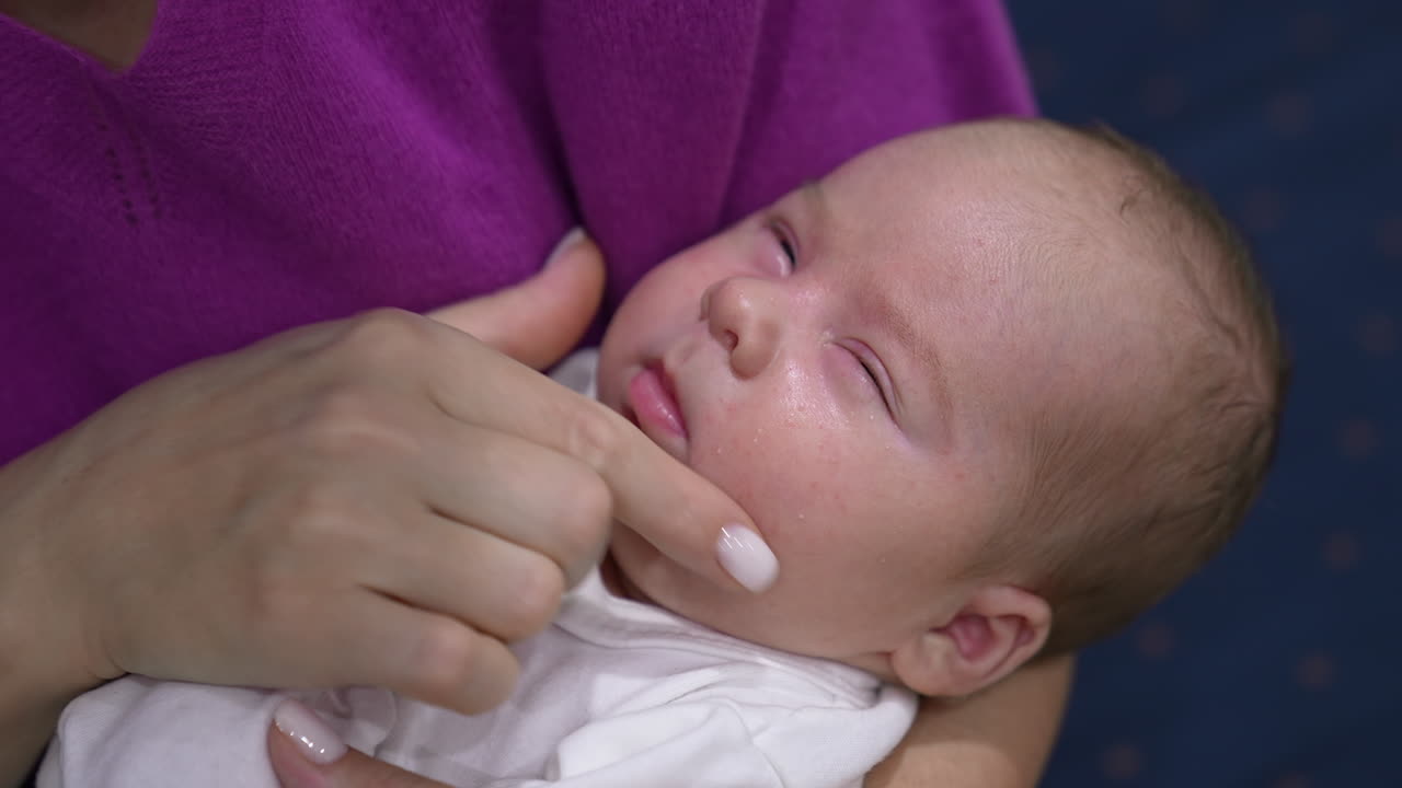 Slowly waking up baby opens and closes his eyes sleepily. Caring mother is pressing baby's cheeks lovingly in attempt to wake him up. Close up.