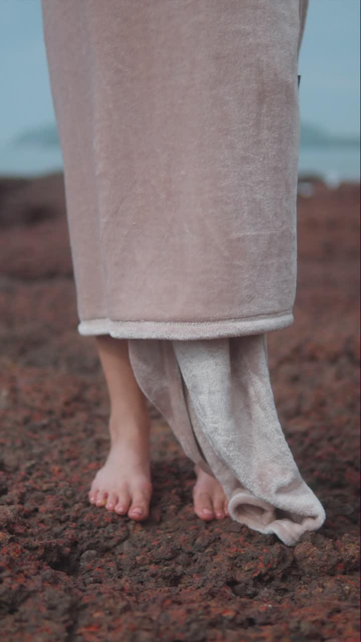 Vertical video of a barefoot woman standing on a rocky shore, wrapped in a soft blanket by the sea