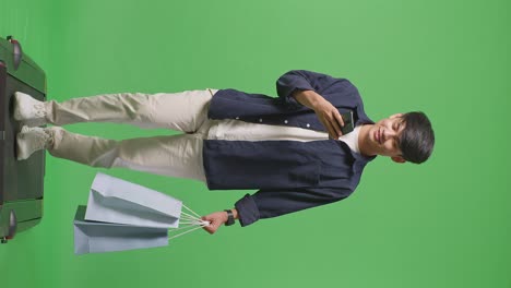 un hombre de compras asiático con bolsas de compras usando un teléfono móvil y sonriendo mientras camina frente a la pantalla verde