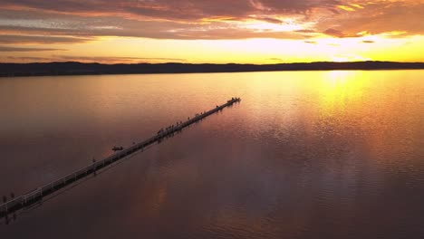 Drone-aerial-view-side-panning-over-paradise-beach-golden-fire-sunset-at-the-Long-Jetty-Wharf-pier-Sydney,-Central-Coast,-Australia