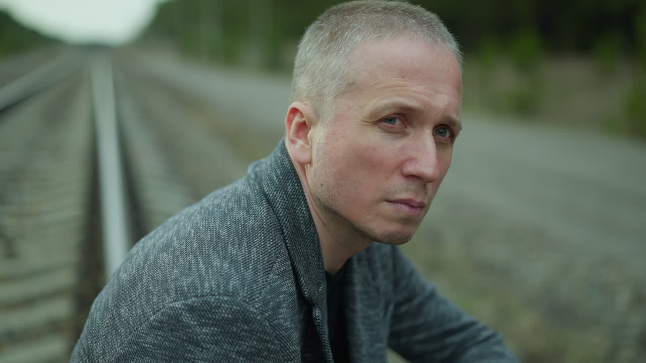 близкий взгляд на задумчивого человека, сидящего у железнодорожного пути, потерянного в глубоком отражении, на заднем плане - лес и электрические столбы