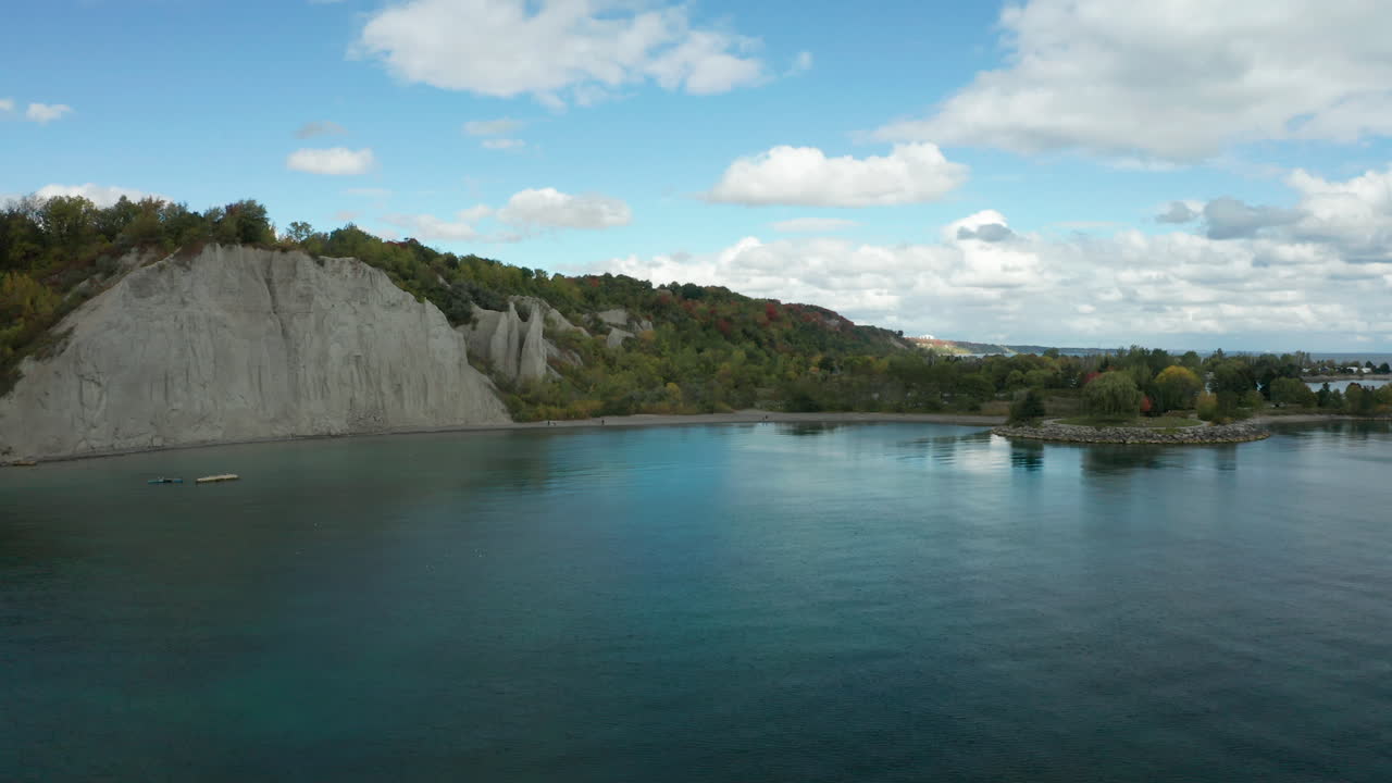 녹색과 주황색 잎이 있는 나무를 드러내기 위해 스카보로 절벽을 향해 날아가는 캐나다 온타리오 호수의 맑은 물 위로 무인 항공기가 발사되었습니다.