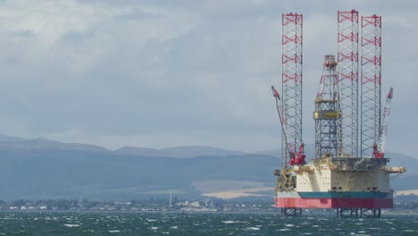 large offshore oil rig platform slowly traverses rough, choppy sea under cloudy daylight, with distant hills and coastline visible in the background. lateral camera pan