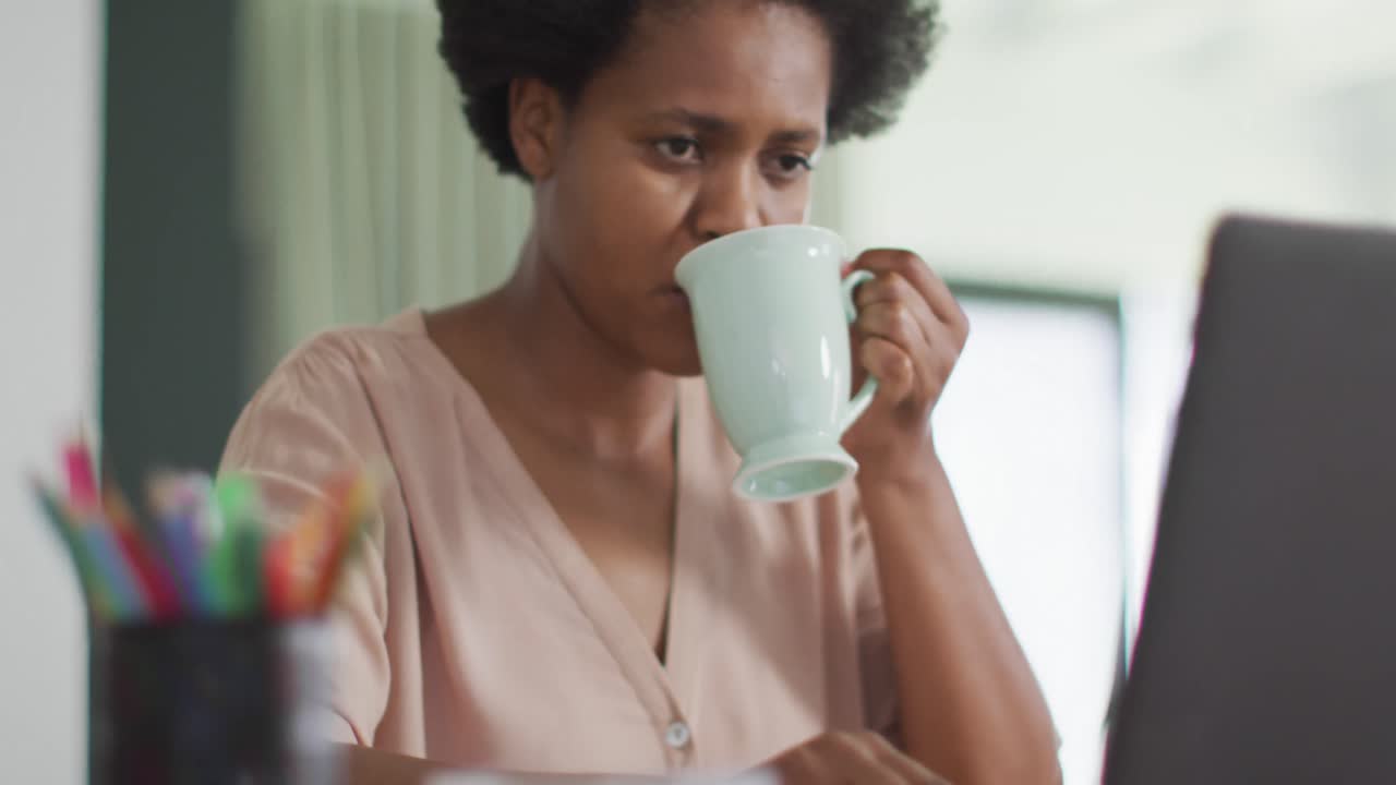 feliz mujer afroamericana sentada en la mesa usando una computadora portátil y bebiendo café