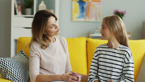 blonde mother holding a cup of coffe and sitting with her teenage daughter on yellow couch at home and having a nice talk