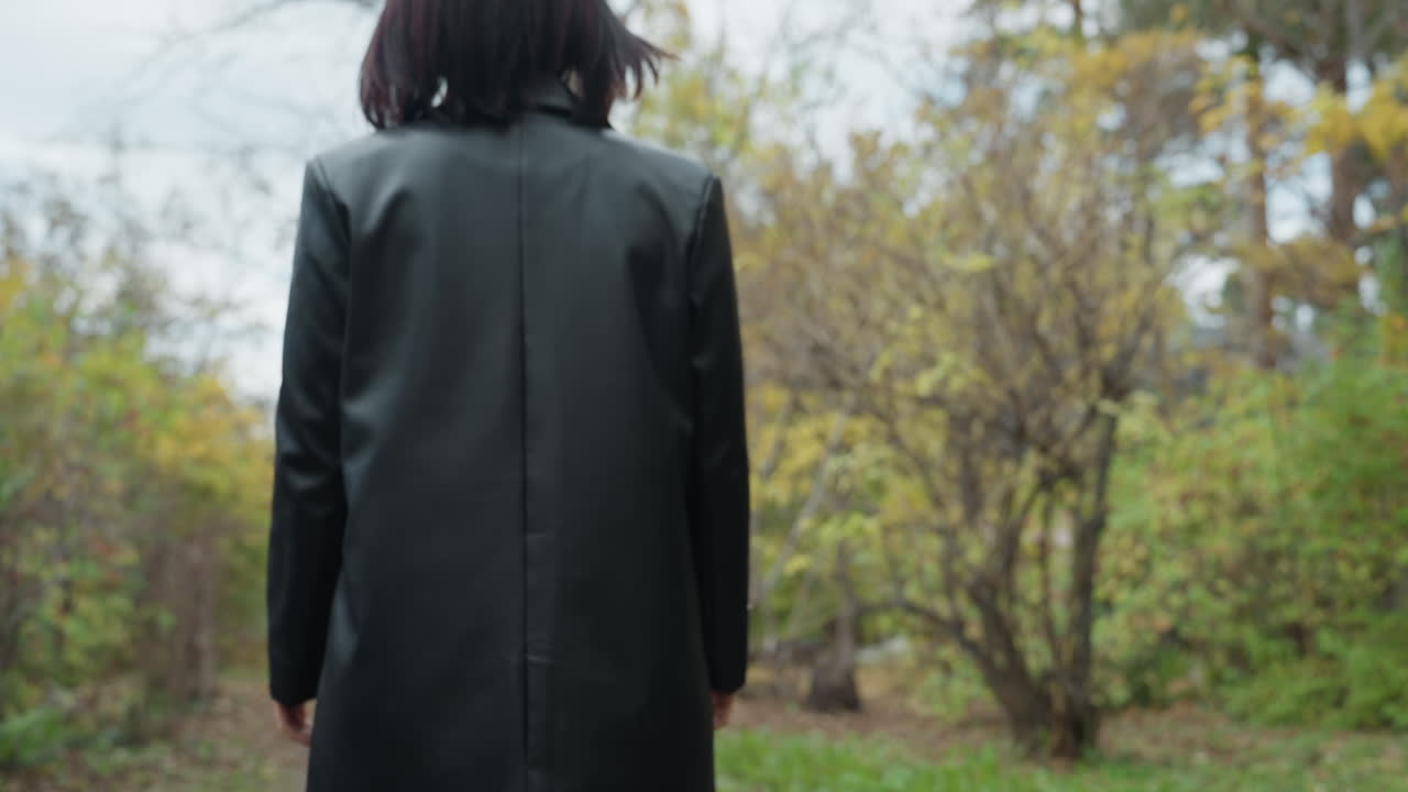 white woman walking in autumn forest, leather jacket and green turtleneck, solitary stroll along leaf covered path, front and back perspectives, reflective expression, overcast sky, crisp air and soft