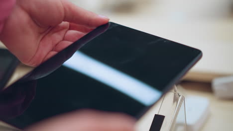 lady picks up a tablet from its stand in an electronics store, connected by an electric cord, she holds it gently, observing the screen with light reflecting off it with the background well lit up