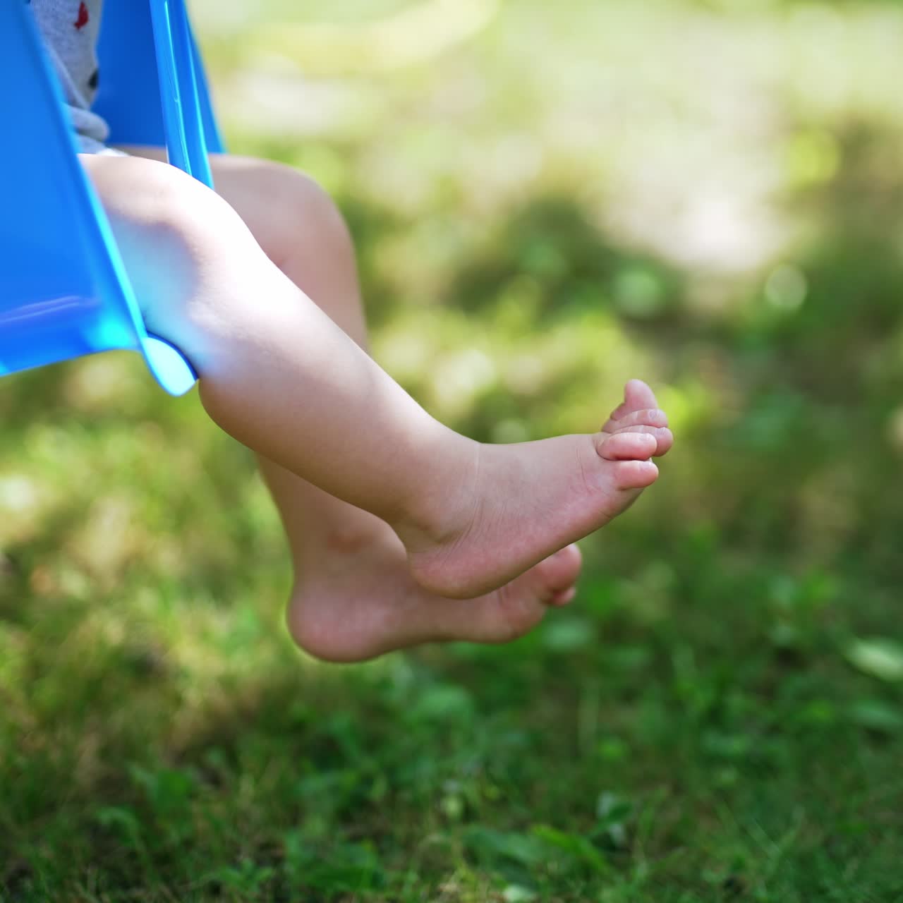 Cute little bare feet of a baby swinging on a blue swing. Happy childhood concept. Baby spending time in the nature. Blurred background