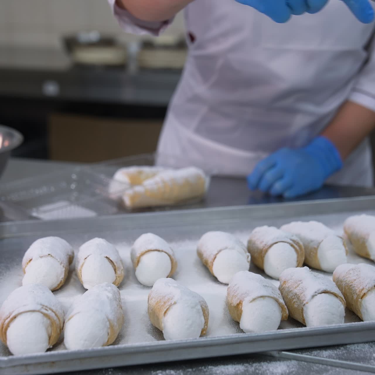 Savory cannoli sweets laid on the metal tray for decoration. Worker covers desserts with sugar powder from a sieve. Close up