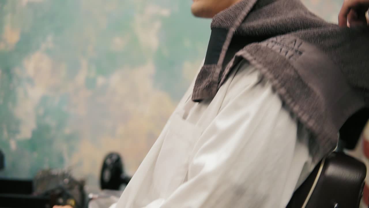 Attractive young man with piercing and tattoo sitting in a chair while bearded barber is preparing him for a haircut. Stylish