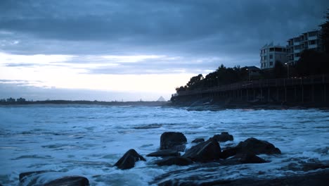 Small-waves-crash-in-front-of-a-raised-wooden-beach-boardwalk-in-the-late-afternoon-as-the-sun-beams-a-blue-tint-into-the-sky-making-the-water-and-its-foam-look-chilly-as-a-mountain-sits-center-frame