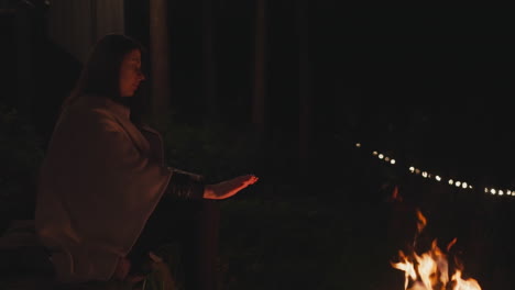 woman sits near fire in front of garlands. lonely woman in blanket warms by campfire in forest with garland lighting. feelings of hopelessness