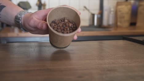 La-Mano-Derrama-Granos-De-Café-Tostados-Marrones-Frescos-Sobre-Una-Mesa-De-Madera