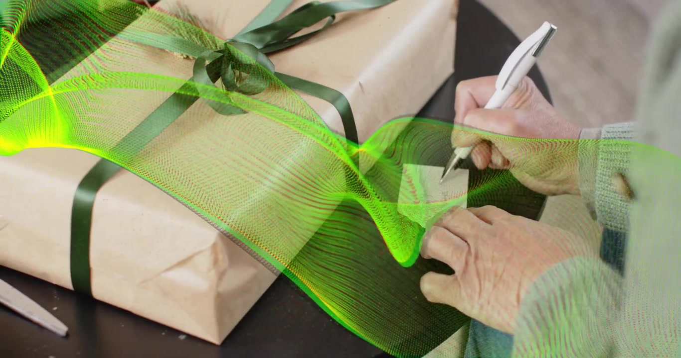 Mãos de uma senhora idosa usando uma caneta para escrever uma etiqueta, amarrando uma fita verde escura em volta de uma caixa bege enquanto faz artesanato.