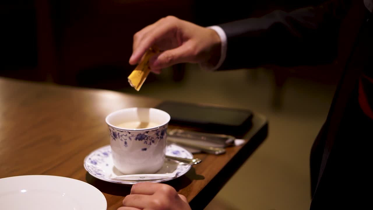 Person hand pouring brown sugar crystals into cup of tea with phone on table