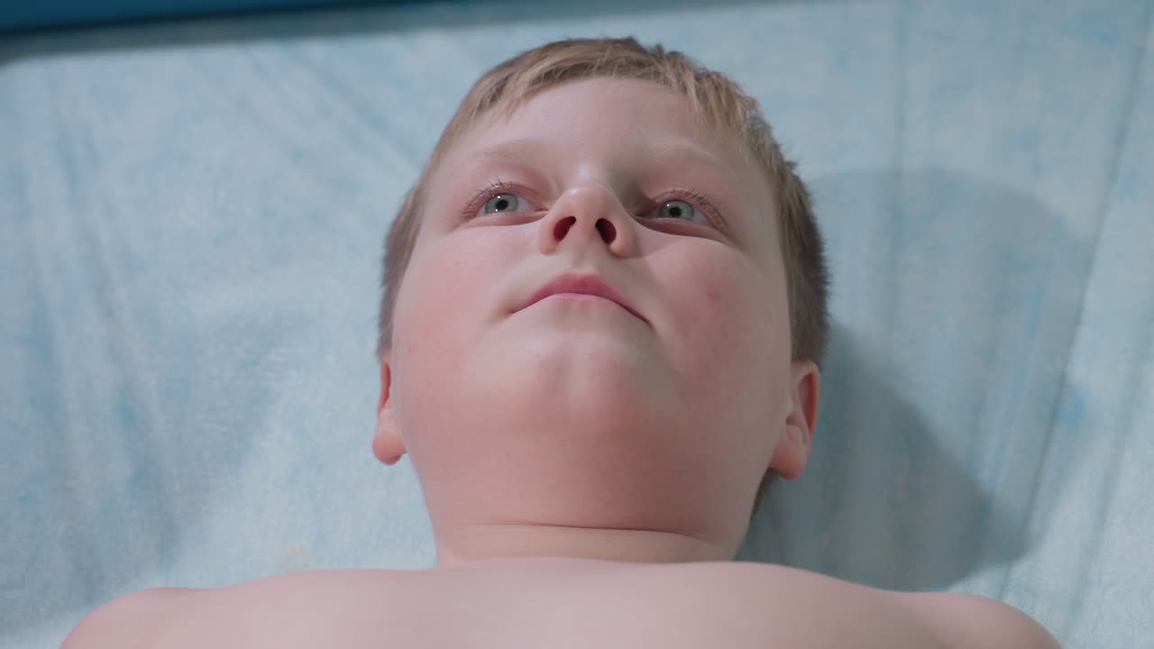Teenage boy lying calmly on lab bed during examination exhales and softly mutters words, expressing a mix of relaxation and curiosity in clinical environment with focus on facial expression
