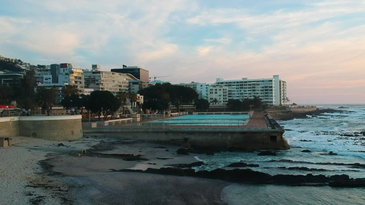 un dron que asciende lentamente por encima de una playa en sea point, ciudad del cabo durante la puesta de sol con piscinas en el fondo