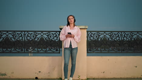 white woman standing outdoors at dusk, leaning against decorative iron fence, wearing headphones, holding phone and looking up thoughtfully, as her clothes flutter gently with wind